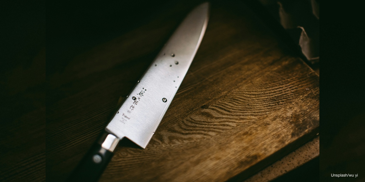 A Japanese Gyuto and a Western chef's knife side by side on a dark stone surface, highlighting the contrasting blade geometry and handle construction of each tradition.