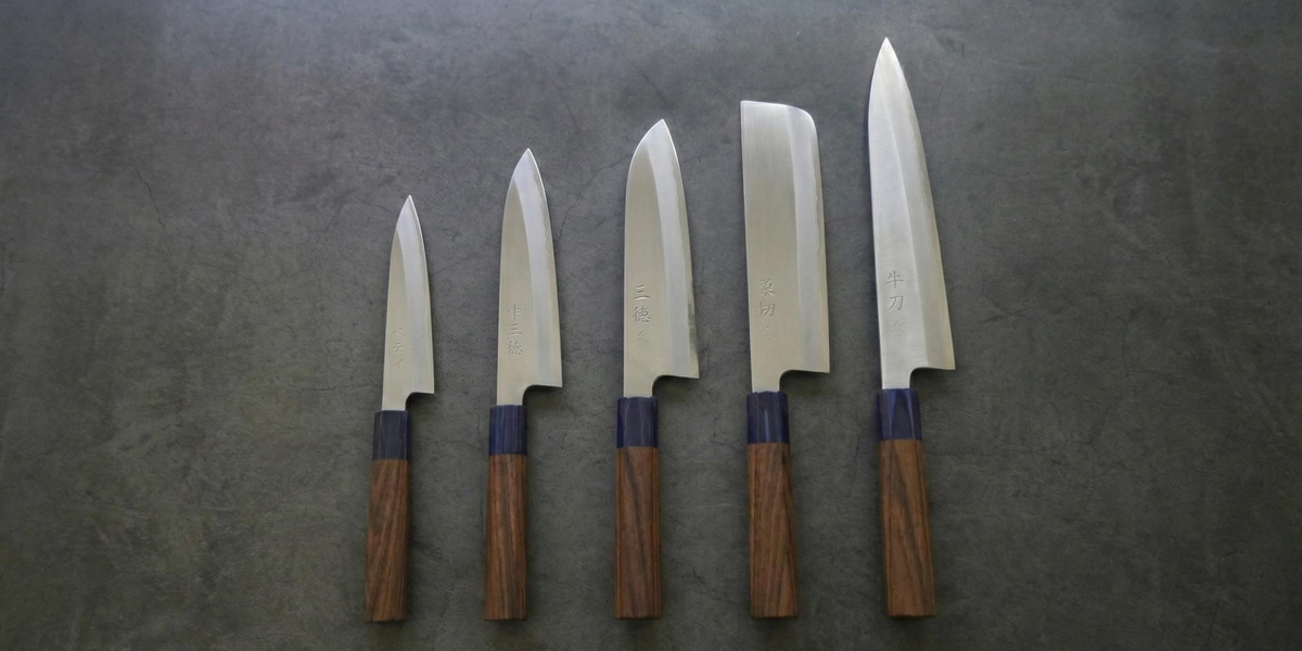 A professional lineup of five traditional Japanese kitchen knives with wooden wa-handles, featuring Petty, Santoku, Nakiri, and Gyuto blades for authentic culinary precision.